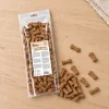 Flat-lay SKU 204603 (Akinu Kosti sušenka se zinkem 1 000 g) — sáček shora na světlém dubovém stole s rozloženými sušenkami-kostičkami. Maketa v Heliosu chybí — reference: BaseLinker čelní foto 1be2e52c + packshot se sušenkami 80011e92 (job 6b286998). 2k 1:1, vybrána v2 ze 2.