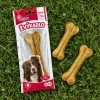 Flat-lay shora na zelené trávě: sáček žvýkadla 204045 (kost z buvolí kůže 16 cm) leží naplocho, vedle dvě volné lisované kosti. Přirozené denní světlo, fotorealistické.