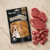 Flat-lay Masíčka Premium Dental 75g: maketa + AI-vygenerovaná fotka syrového hovězího masa (mix plátků a kostiček) jako surovina-referenceo; travertinová deska, evergreen kuchyňský mood. Kompoziční reference pro řadu nebyla v Content DB nalezena (content_list_assets kind=obrazek_reference/subkind=kompozicni_reference/tags flat_lay_kompozice+akinu_masicka_premium vrátil 0 výsledků) — generováno jen podle textového popisu kompozice z Kroku 9.