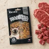 Flat-lay Akinu Masíčka Premium Heart & Eyes pro kočky 50 g (282038) — sáček mírně mimo střed na matném travertinu, syrové hovězí (plátky + kostičky) asymetricky v jednom rohu. Varianta f1 ze 4. Surovinová reference = AI generace syrového hovězího (job 15945286); kompoziční reference řady v Content DB nenalezena.