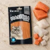 Flat-lay Akinu Masíčka Premium Losos s treskou mini proužky pro kočky 50 g (282056) — sáček mírně mimo střed na matném travertinu, syrový losos a treska asymetricky v jednom rohu. Varianta f1 ze 4. Surovinová reference = AI generace syrového lososa a tresky (job 8a636129); kompoziční reference řady v Content DB nenalezena.