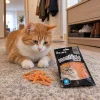 Lifestyle Akinu Masíčka Premium Losos s treskou mini proužky pro kočky 50 g (282056) — zrzavo-bílá kočka klidně leží na koberečku v běžné české předsíni a kouká na pamlsky před sebou, vedle leží sáček; teplé podvečerní světlo. Varianta b1 ze 2.